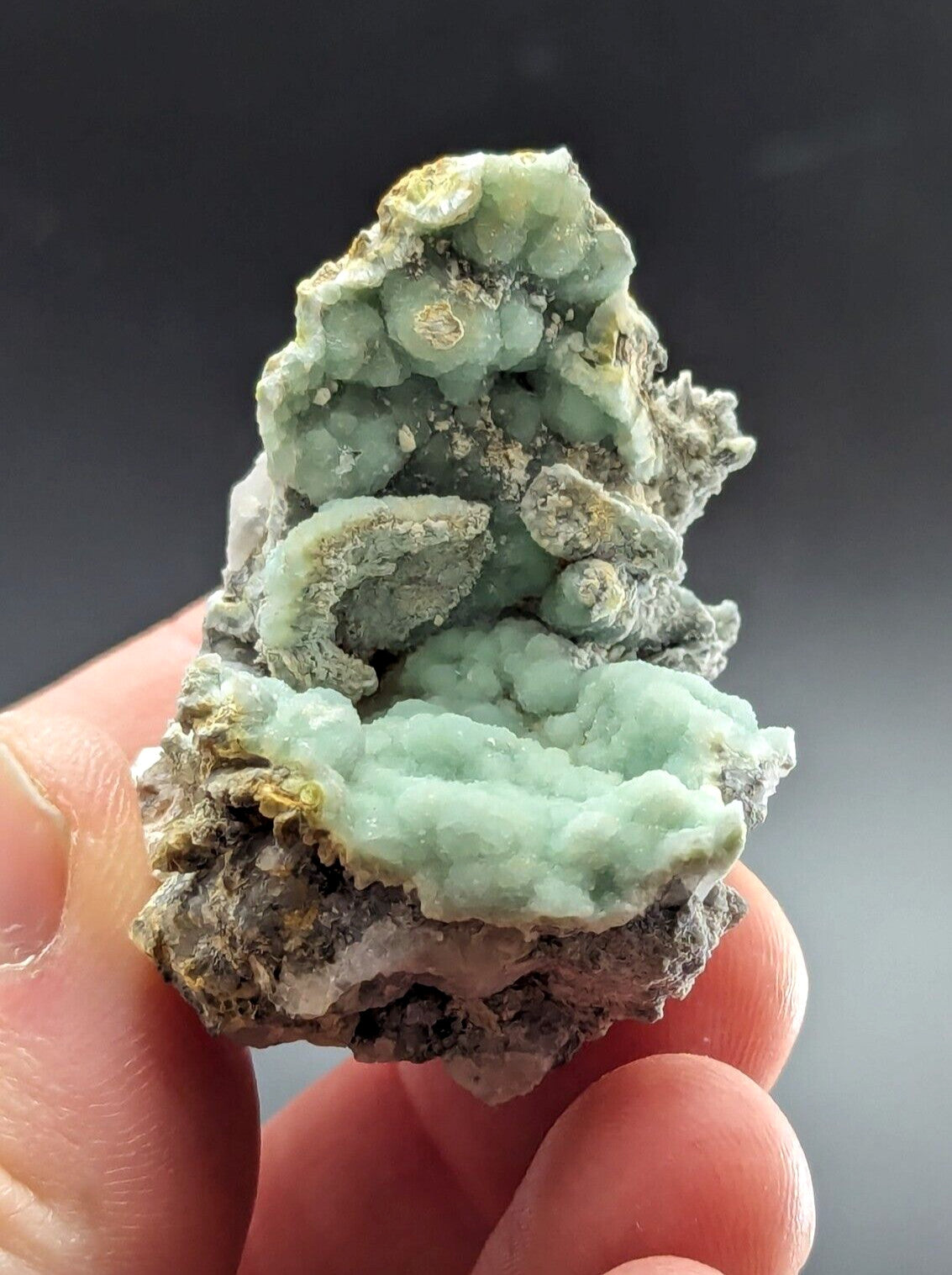 A hand holds a light green, crystalline mineral specimen with rough, textured surfaces and patches of yellowish material.