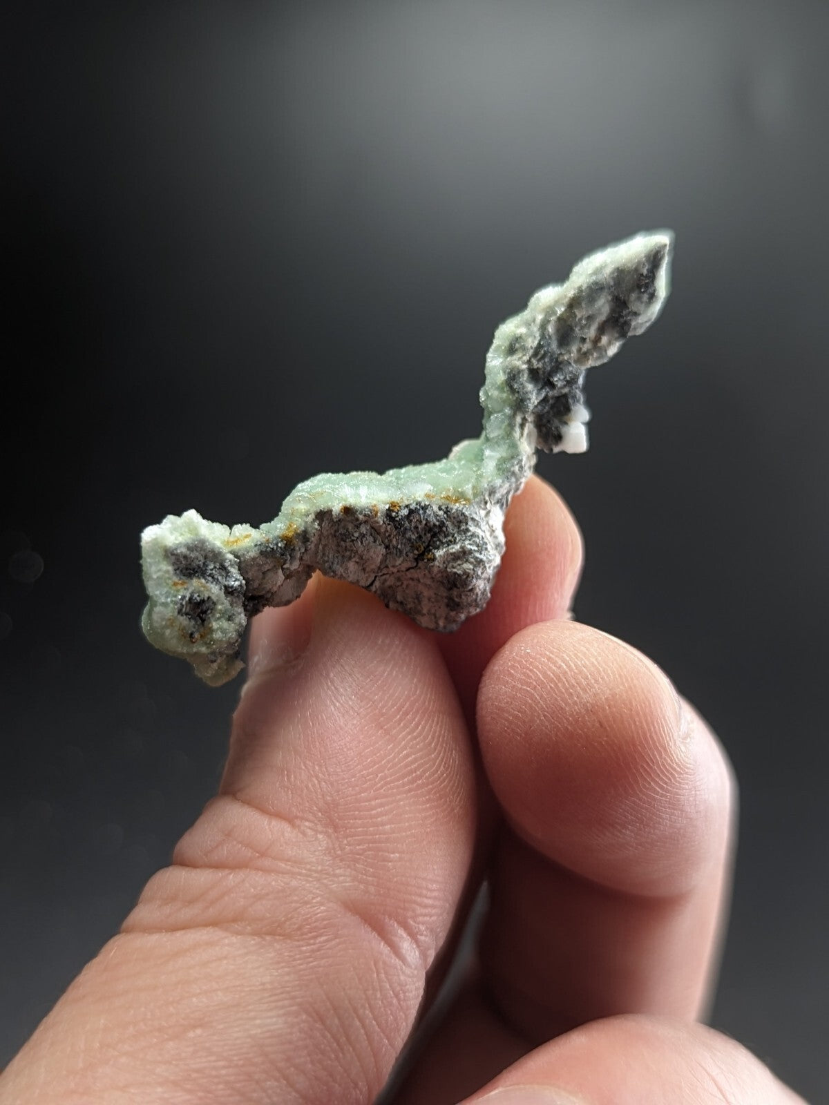 A delicate, translucent green mineral specimen with a rough, crystalline texture and dark gray veining.