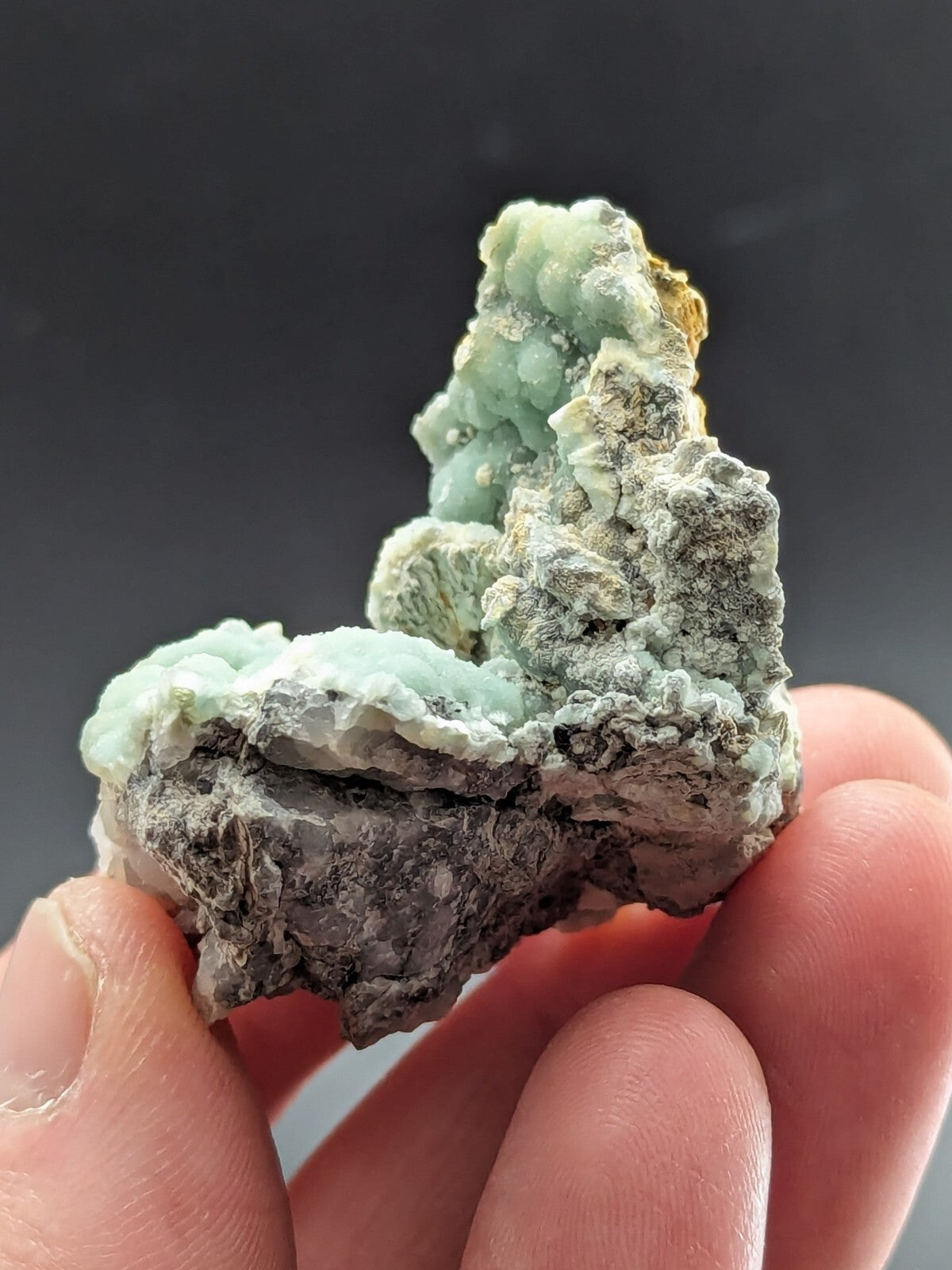 A hand holds a natural mineral specimen featuring light green, crystalline formations atop a darker gray rock base with subtle golden accents.