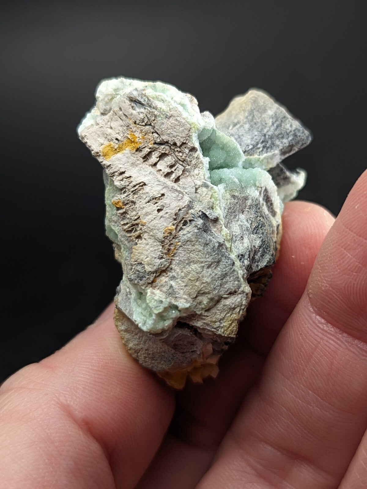 A hand holds a rough, irregularly shaped mineral specimen featuring translucent light green crystals intermingled with grayish rock and patches of yellow-orange oxidation.