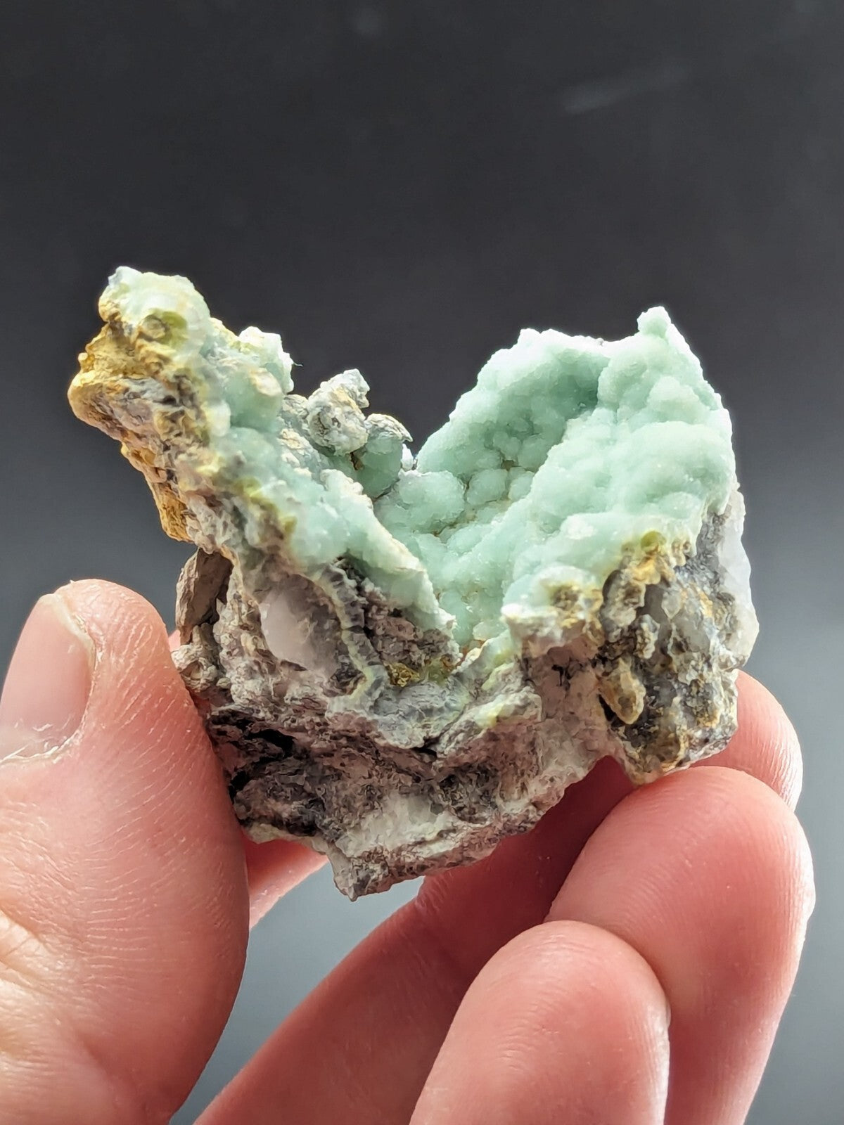 A hand holds a natural mineral specimen featuring vibrant light green, textured crystals with patches of yellow and gray matrix.