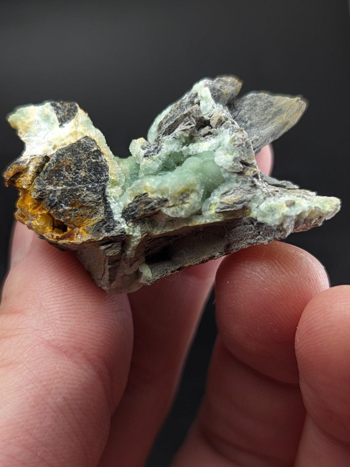 A hand holds a rough, irregularly shaped mineral specimen featuring patches of translucent light green crystals embedded in dark gray and yellowish rock.