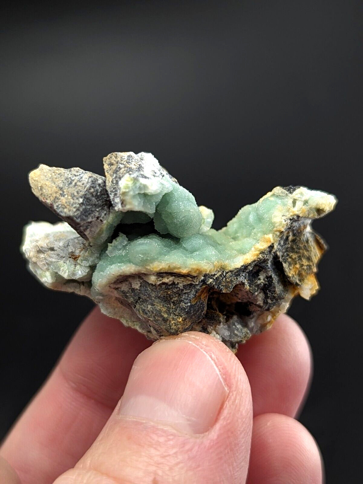 A hand holds a natural mineral specimen featuring vibrant green chrysocolla crystals growing from dark, rugged matrix rock.