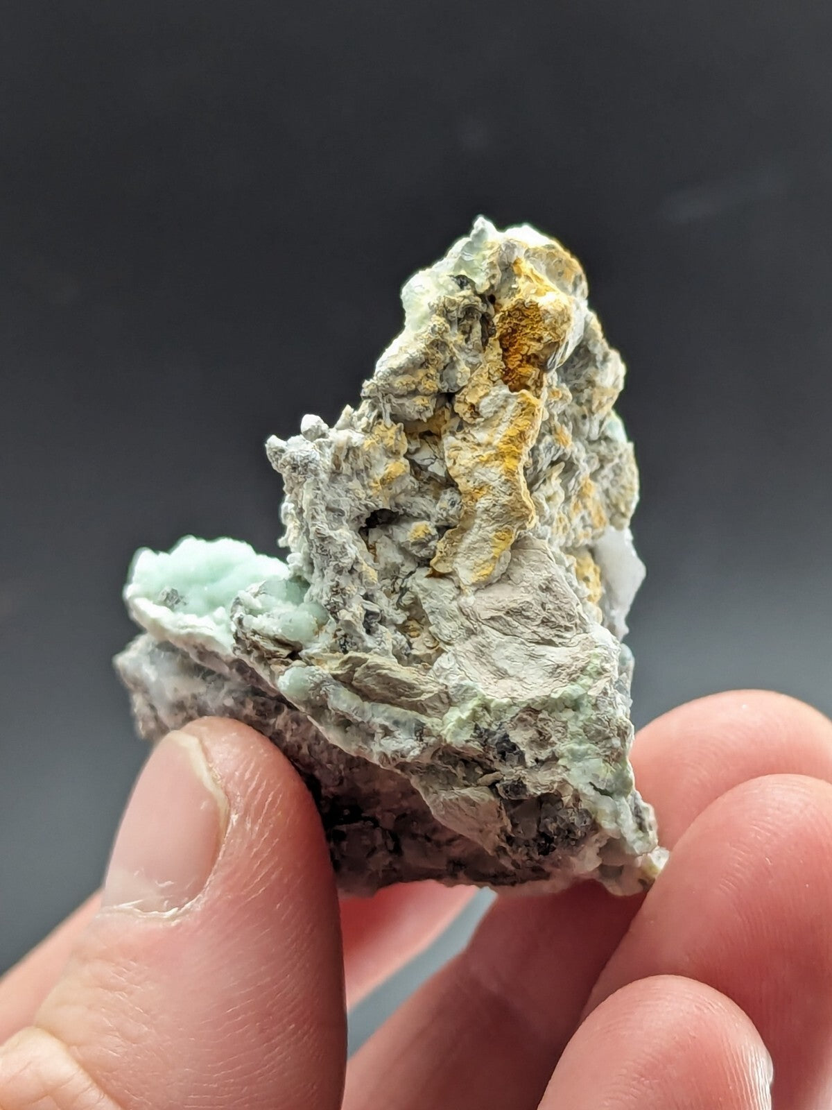 A hand holds a rough, irregularly shaped mineral specimen featuring patches of pale green and yellowish-orange crystals embedded in a gray matrix.