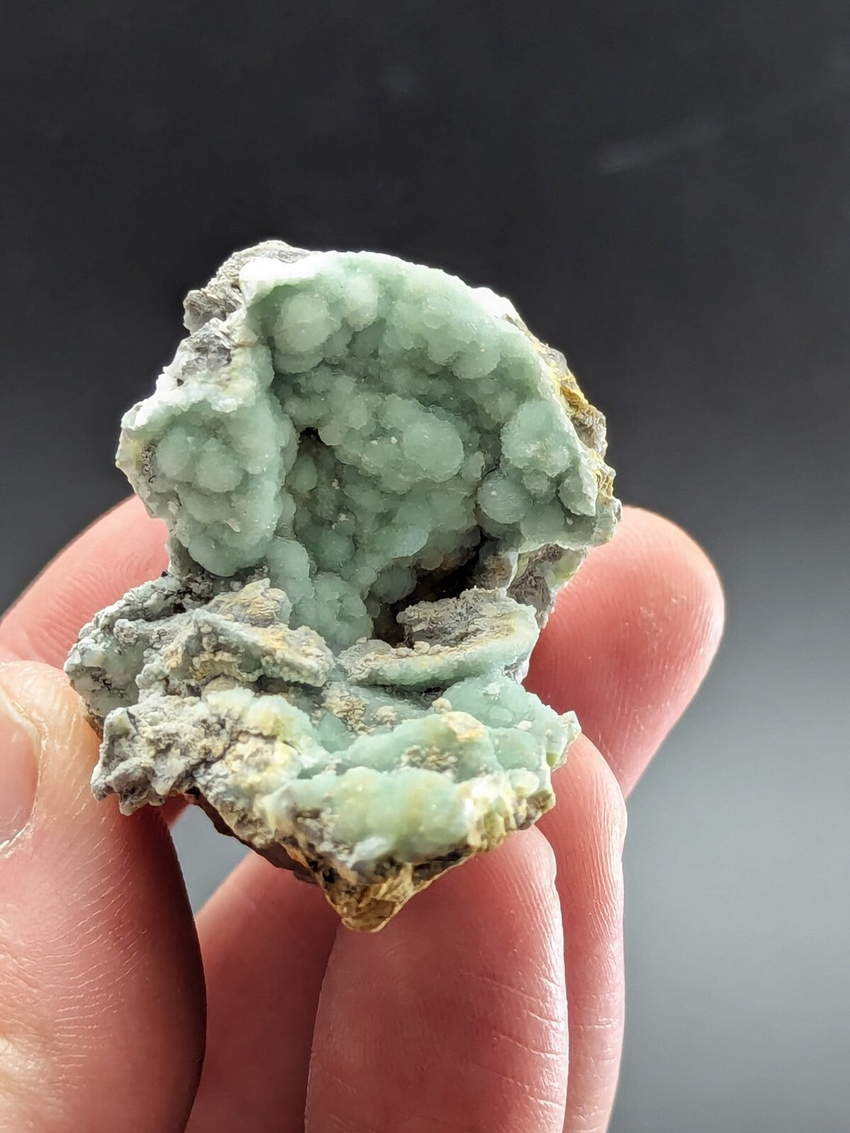 A hand holds a light green, crystalline mineral specimen with rounded, bubble-like formations and patches of grayish matrix.