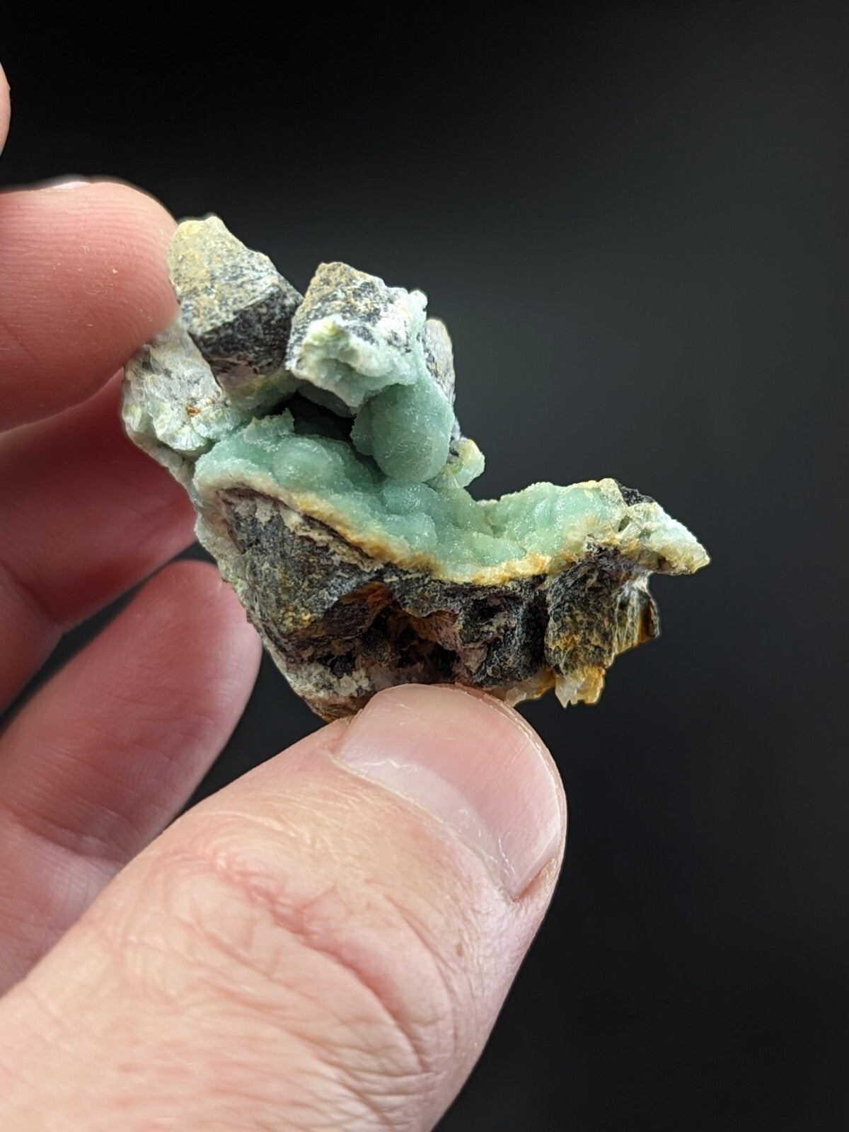 A hand holds a natural mineral specimen featuring vibrant green, bumpy crystals growing from dark gray and brown rock.