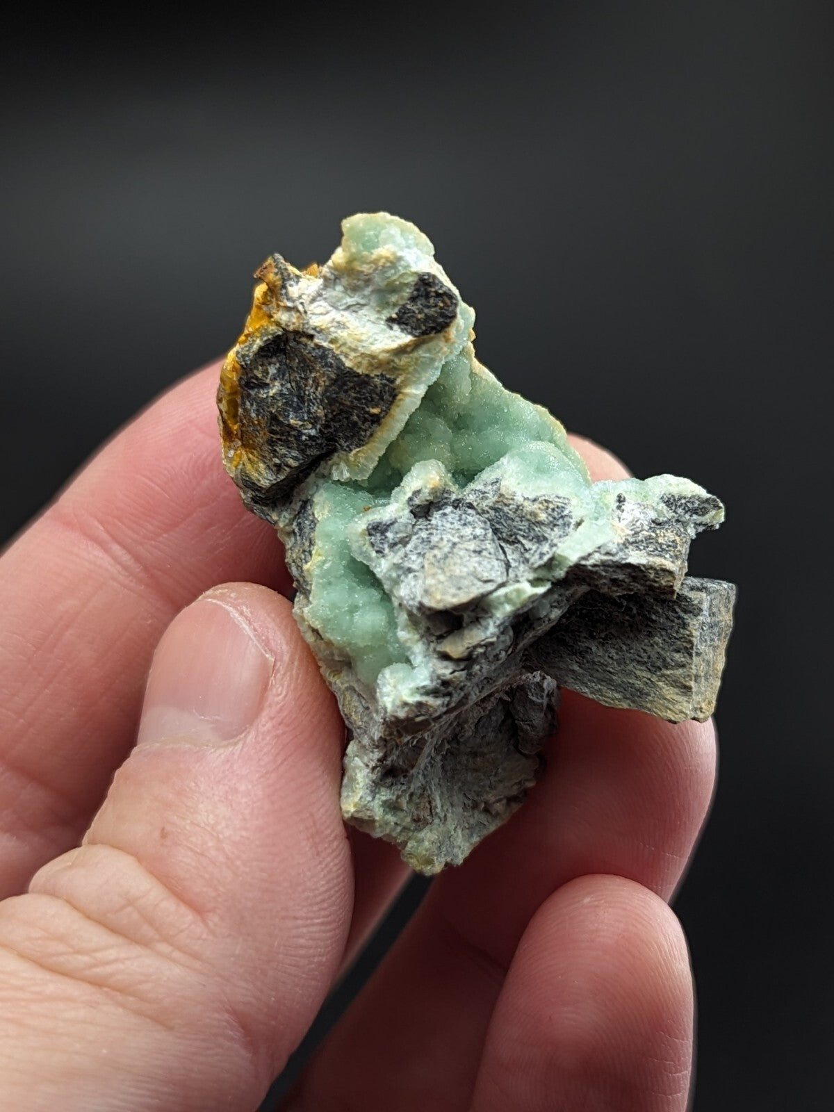 A rough, irregularly shaped piece of greenish-blue mineral with patches of dark gray and yellow-orange matrix.