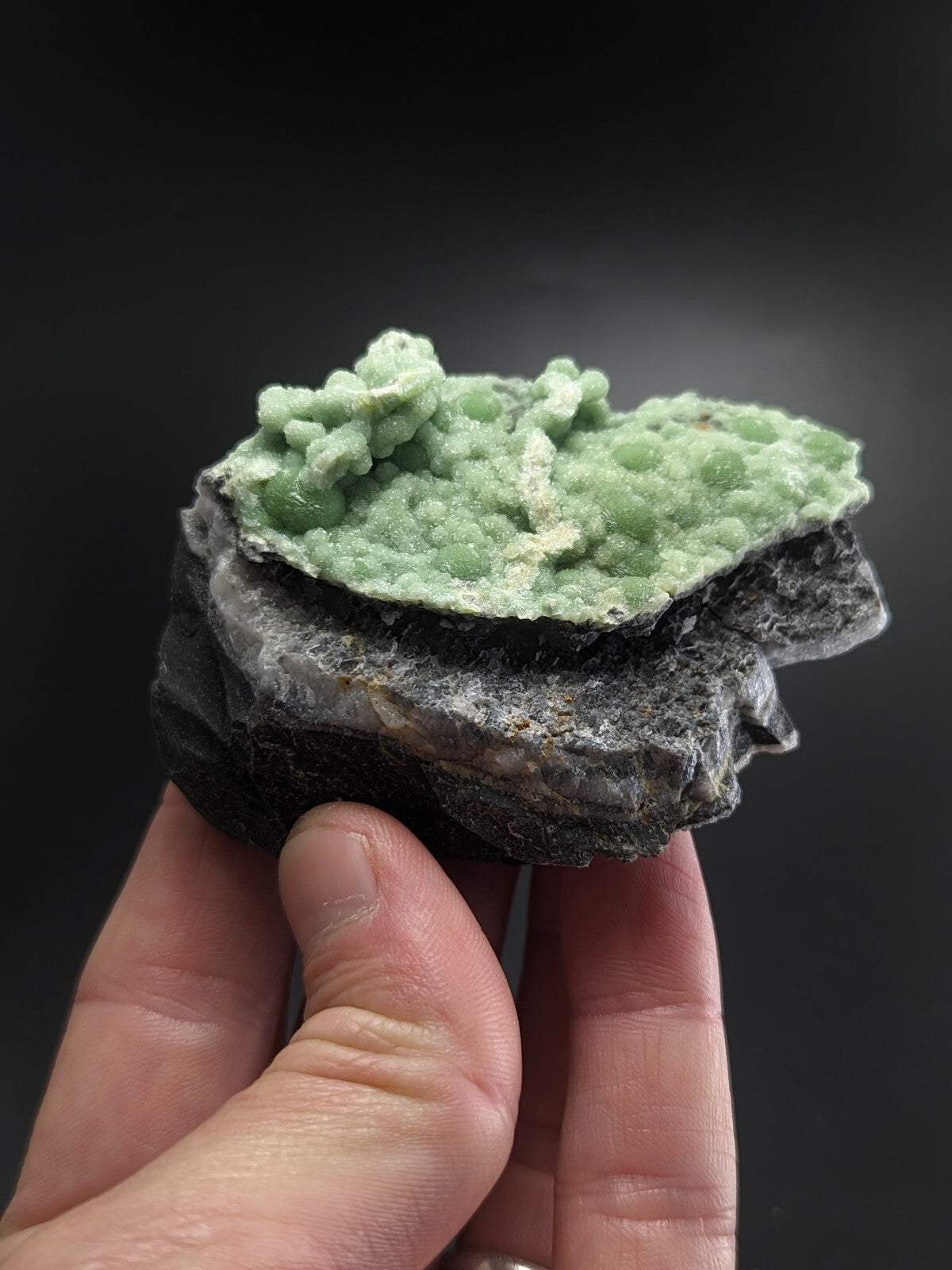 A hand holds a geological specimen featuring vibrant green, crystalline formations atop a dark gray rock base.
