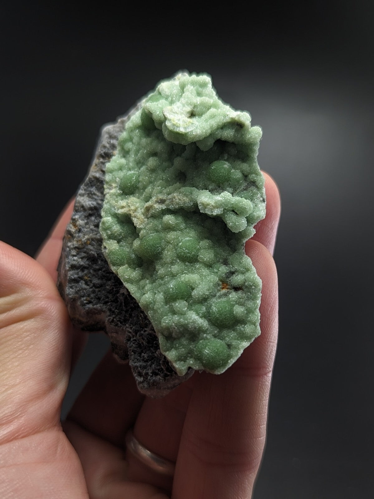 A hand holds a light green, bumpy mineral specimen with a rough texture and some grayish matrix.