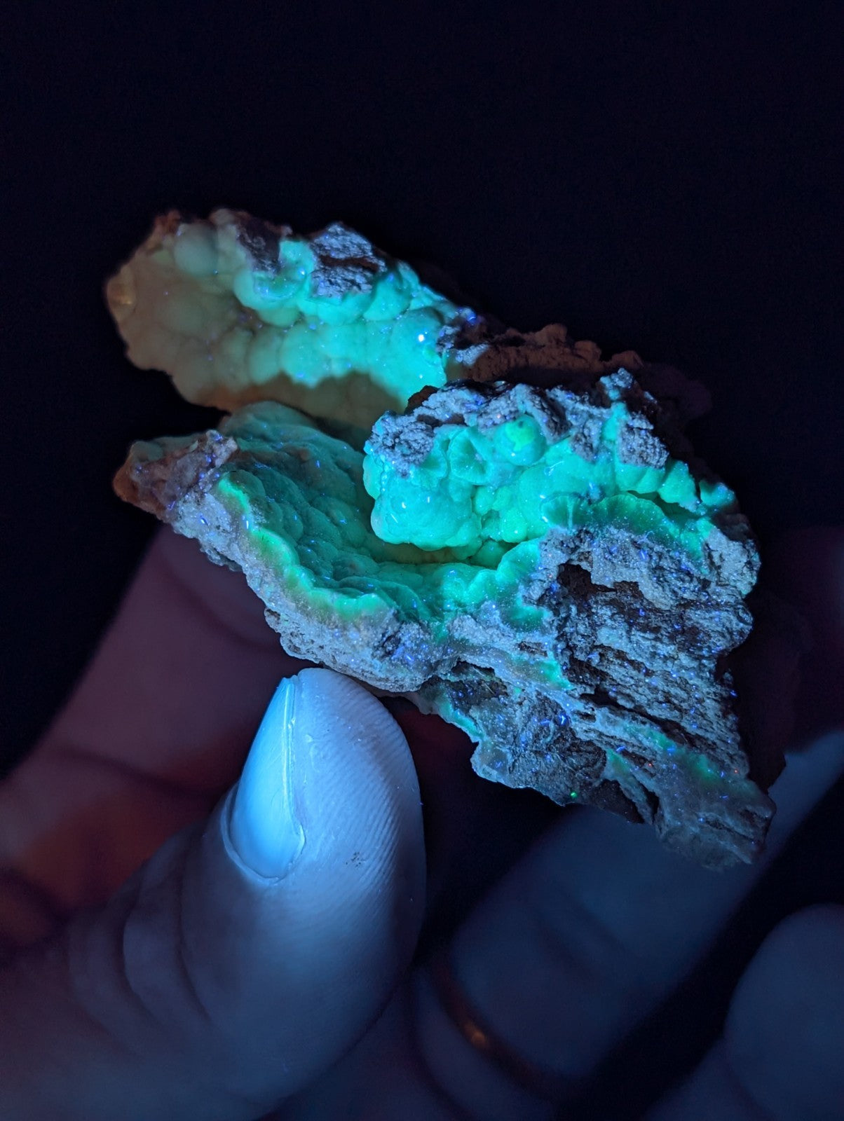 A hand holds a glowing green fluorescent mineral specimen with rough, textured surfaces and earthy brownish outer layers.