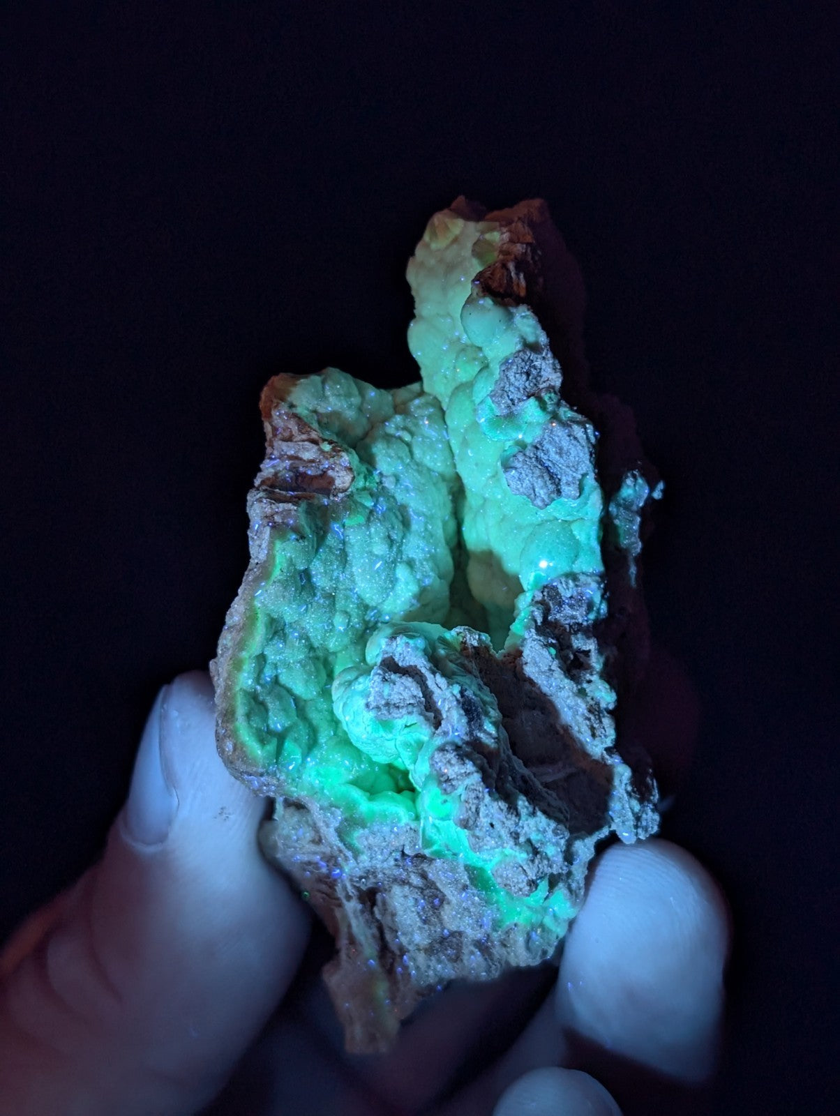 A hand holds a glowing green mineral specimen with rough, textured surfaces and patches of darker earthy tones.