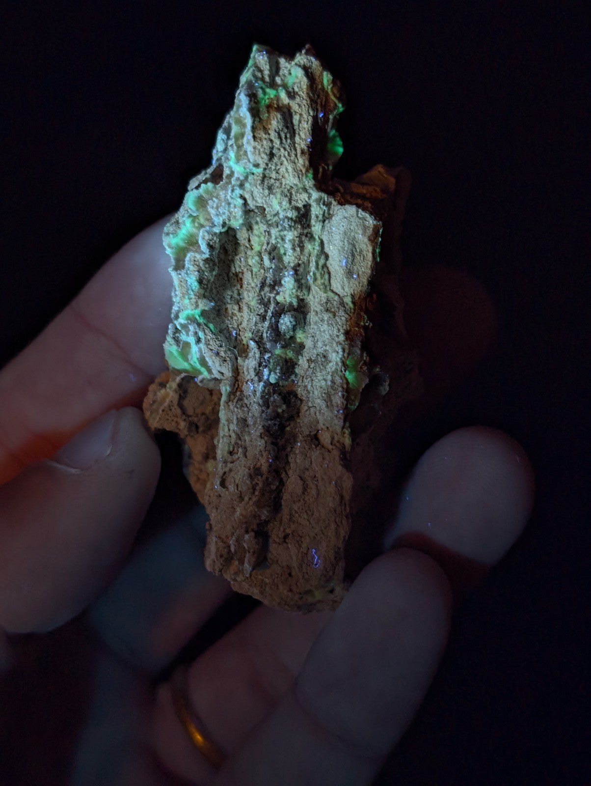 A hand holds a rough, irregularly shaped mineral specimen with patches of glowing green fluorescence against its earthy brown and tan matrix.