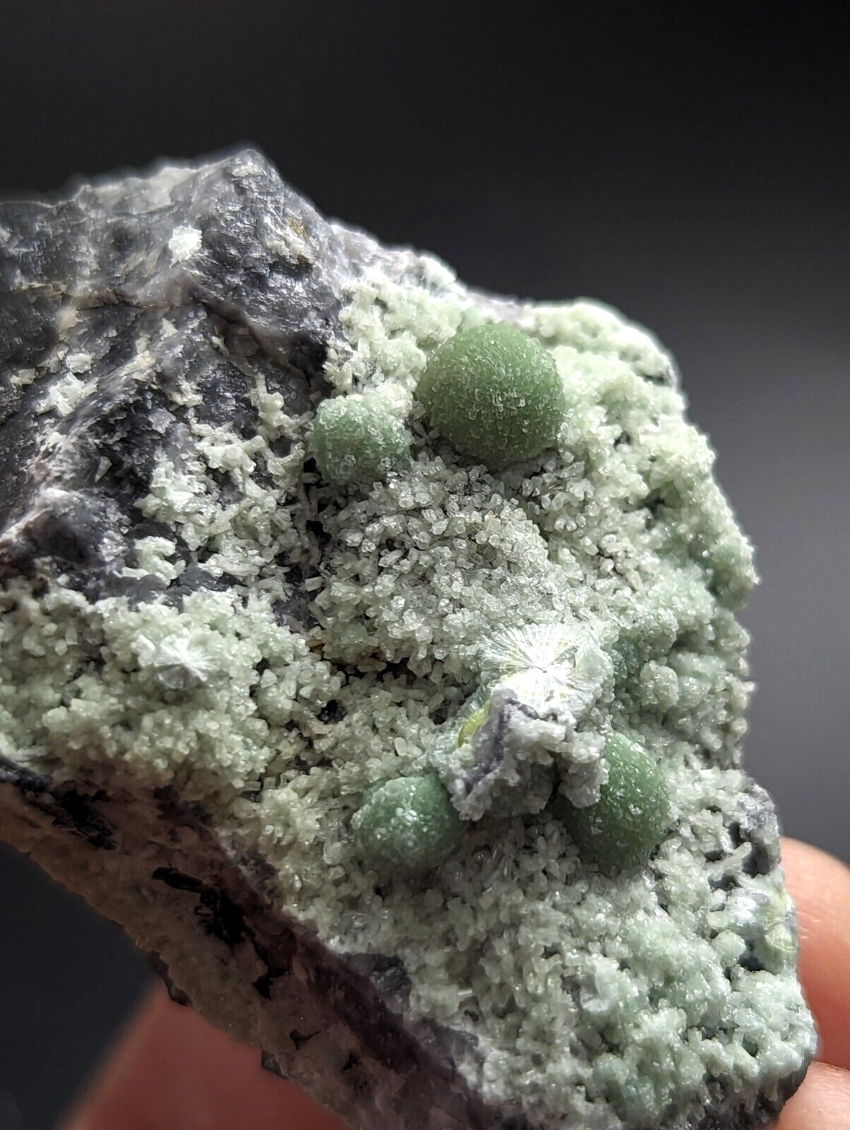 A cluster of vibrant green, spherical crystals grows from a rough, grayish rock matrix.