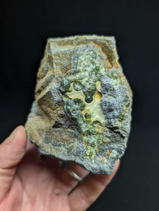 A hand holds a rough, irregularly shaped rock with a grayish base and patches of green mineral growth featuring small, round, translucent crystals.