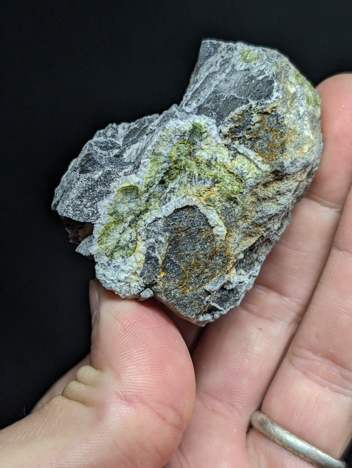 A hand holds a rough, irregularly shaped rock with grayish-white base material and patches of green and yellow mineral growth.