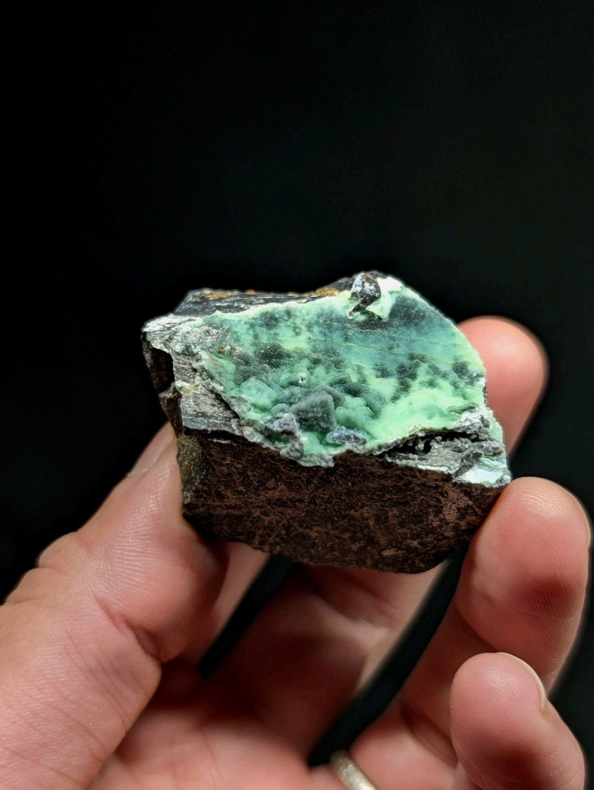 A hand holds a rough, irregularly shaped mineral specimen featuring a vibrant green patina over dark gray rock.