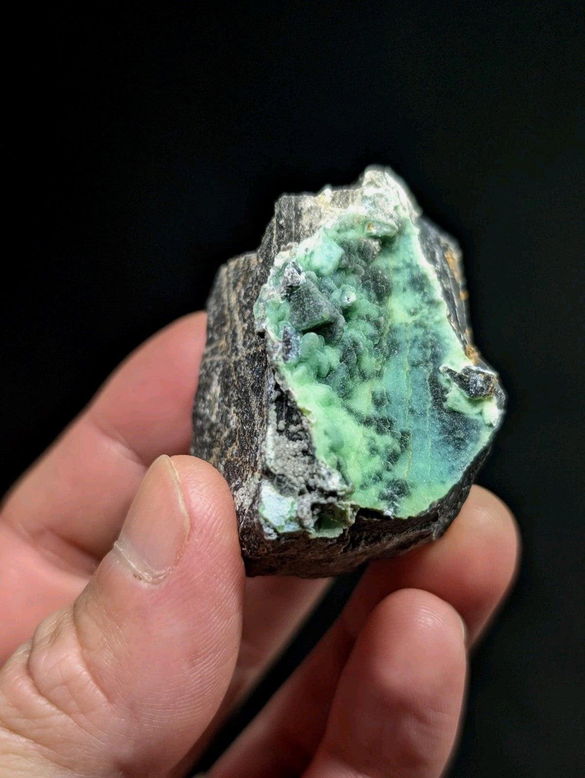 A hand holds a rough, irregularly shaped piece of green mineral with dark gray matrix.