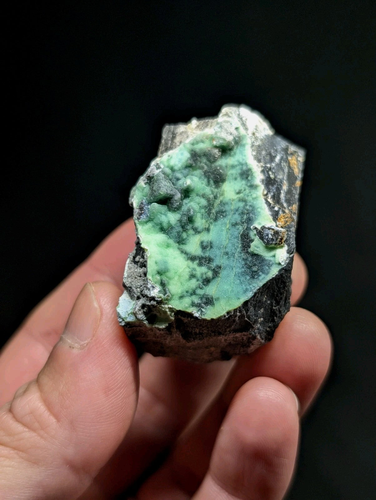 A hand holds a rough, irregularly shaped mineral specimen featuring vibrant green veining against dark gray and black rock matrix.