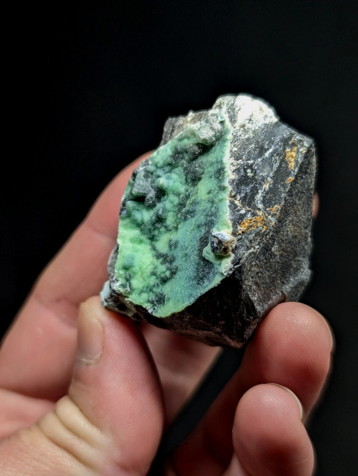 A hand holds a rough, irregularly shaped mineral specimen featuring vibrant green veining against a dark gray matrix with subtle golden flecks.
