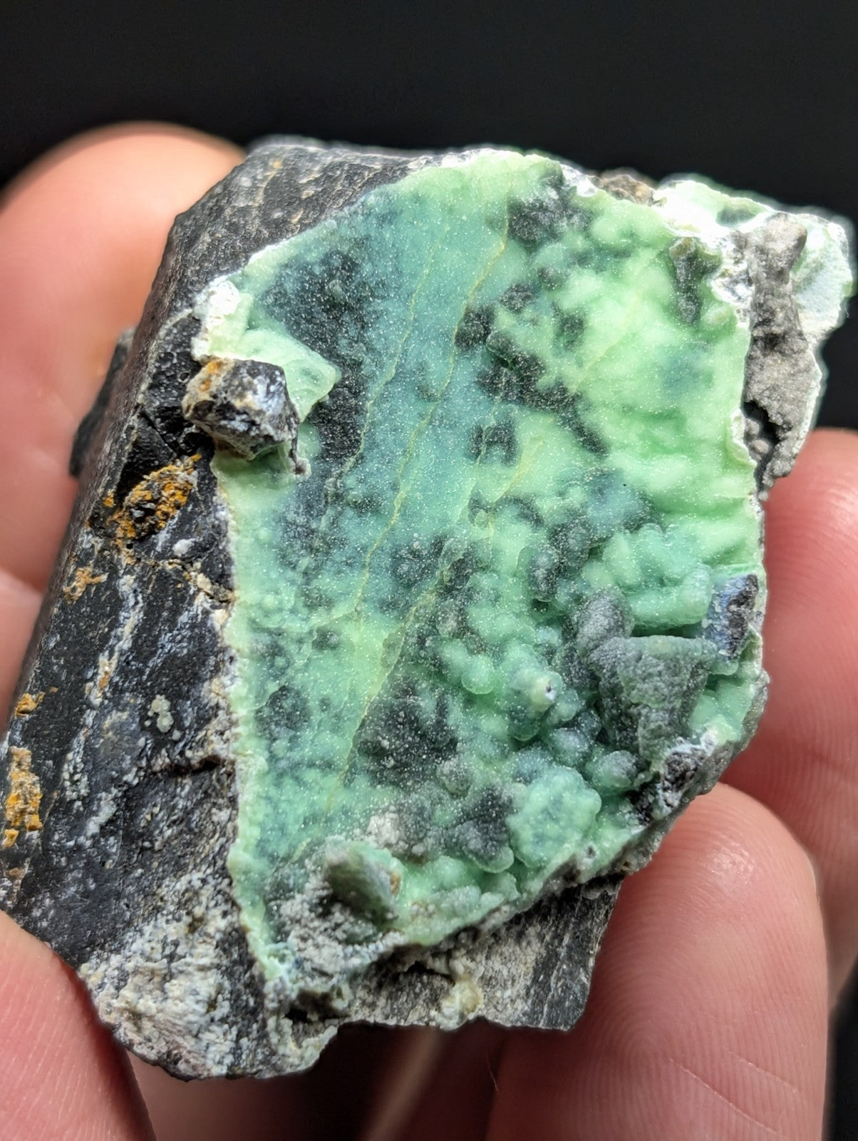 A rough, irregularly shaped mineral specimen featuring a vibrant green crystalline formation set against a dark gray matrix with patches of yellow and white.