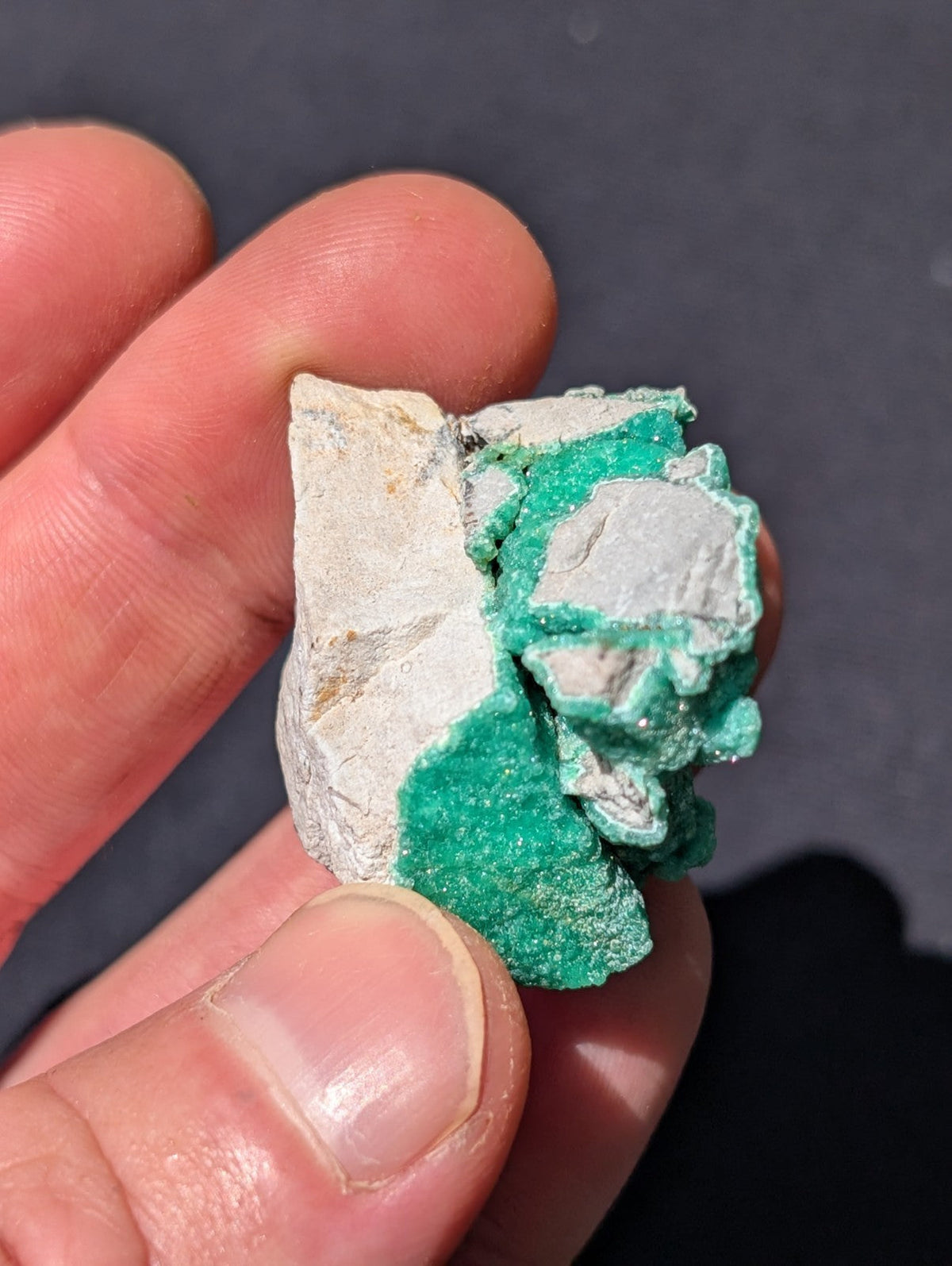 A hand holds a rough, irregularly shaped mineral specimen with a white base and vibrant green crystalline formations.