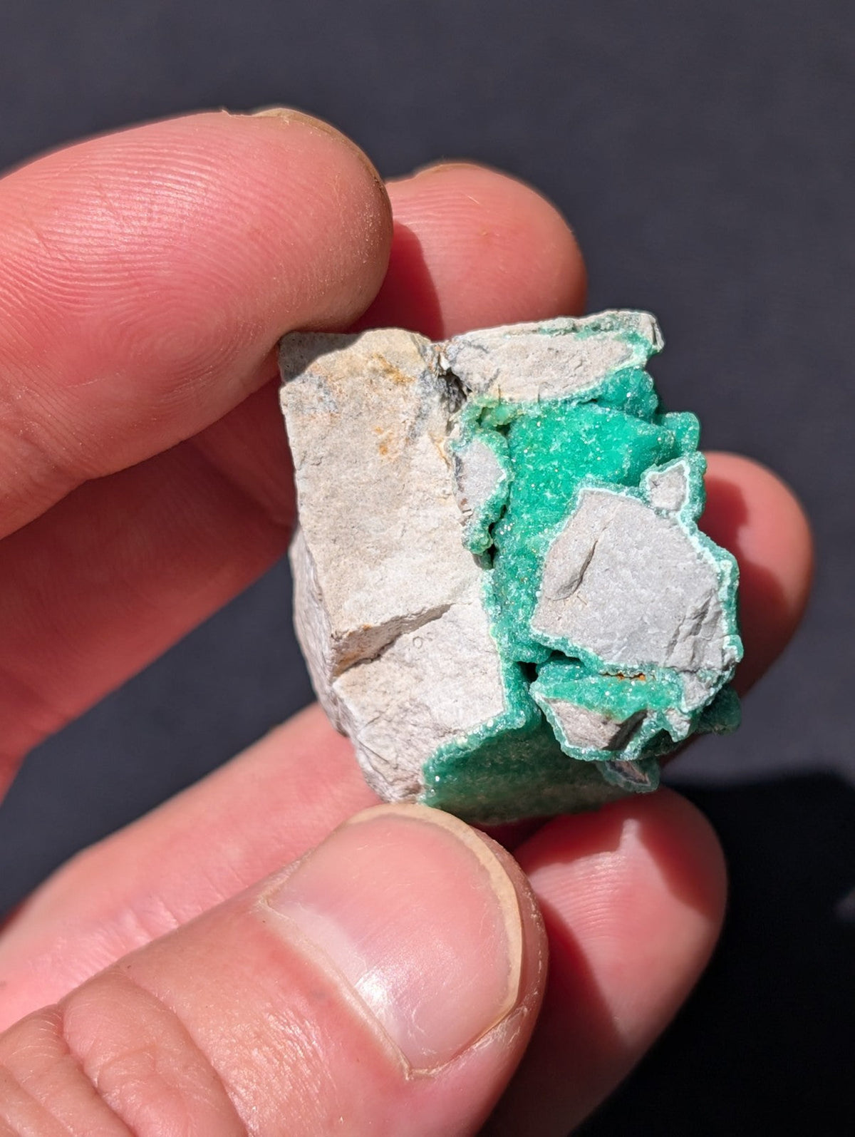 A hand holds a rough, irregularly shaped mineral specimen featuring vibrant green crystals embedded within a lighter gray matrix.