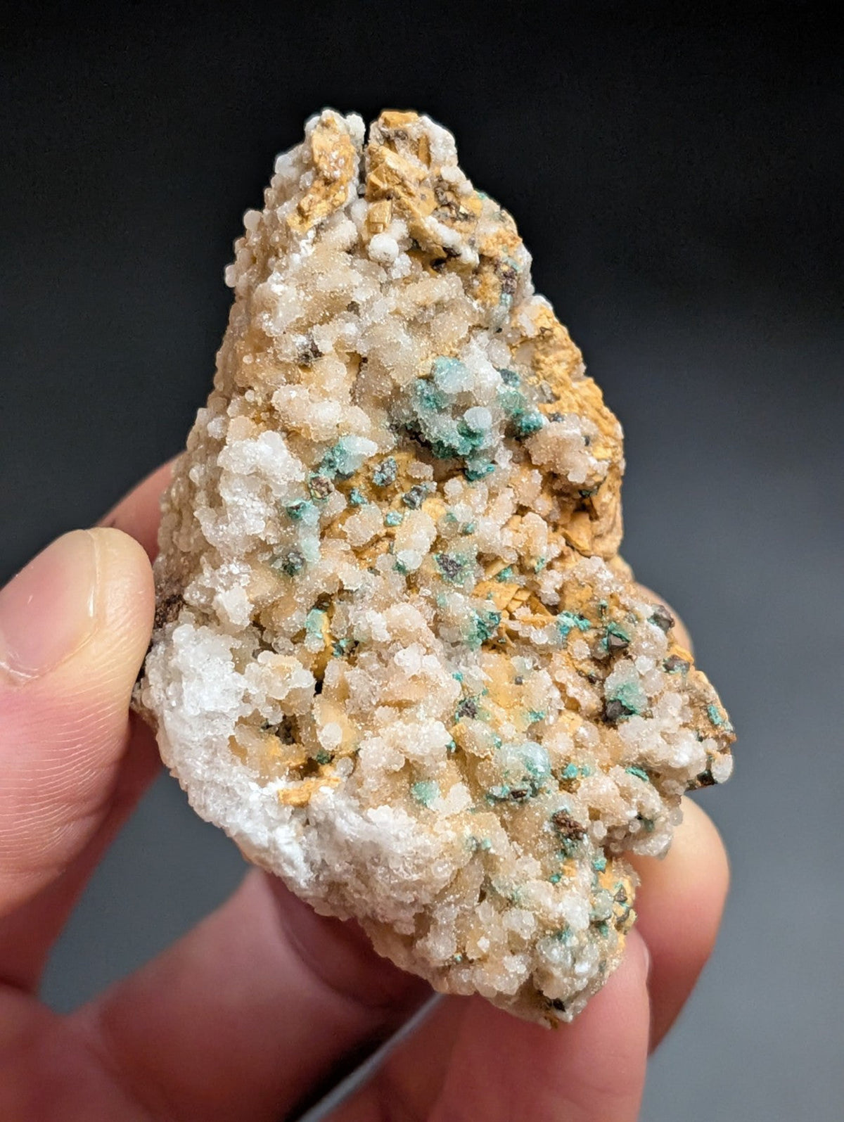 A hand holds a naturally occurring mineral specimen featuring a textured surface with crystalline formations in white, pale yellow, and patches of vibrant green.
