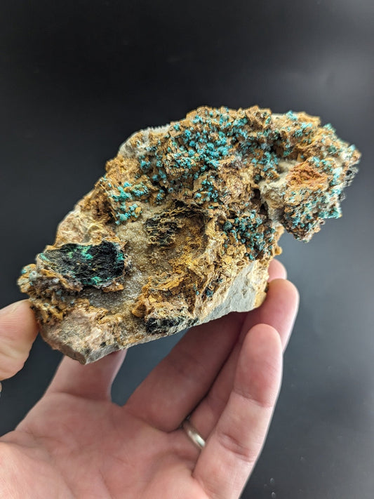 A hand holds a rough, irregularly shaped mineral specimen featuring vibrant turquoise and green crystalline formations over a base of earthy brown and gray rock.