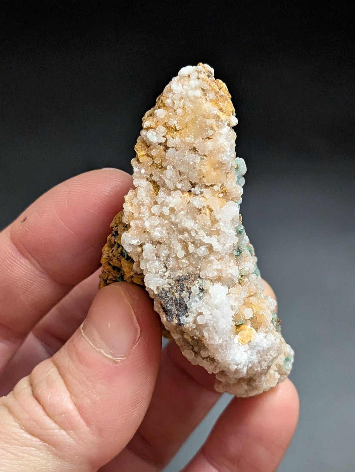 A hand holds a naturally occurring mineral specimen featuring a textured surface with white, crystalline formations overlying yellowish and greenish matrix rock.
