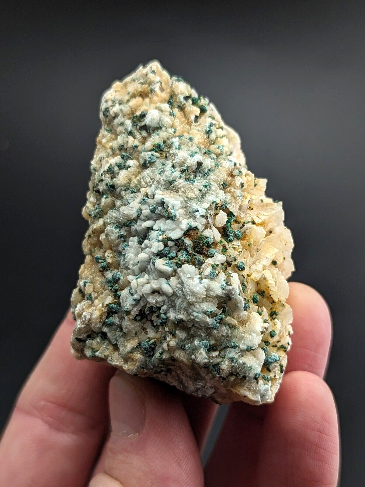 A hand holds a rough, irregularly shaped mineral specimen featuring patches of creamy white and vibrant green crystalline formations against a pale beige matrix.
