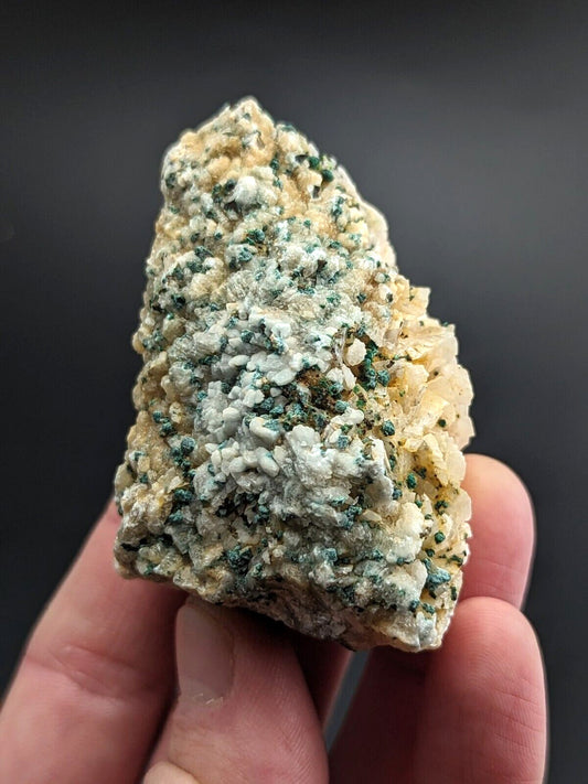 A hand holds a rough, irregularly shaped mineral specimen featuring patches of creamy white and vibrant green crystalline formations against a pale beige matrix.