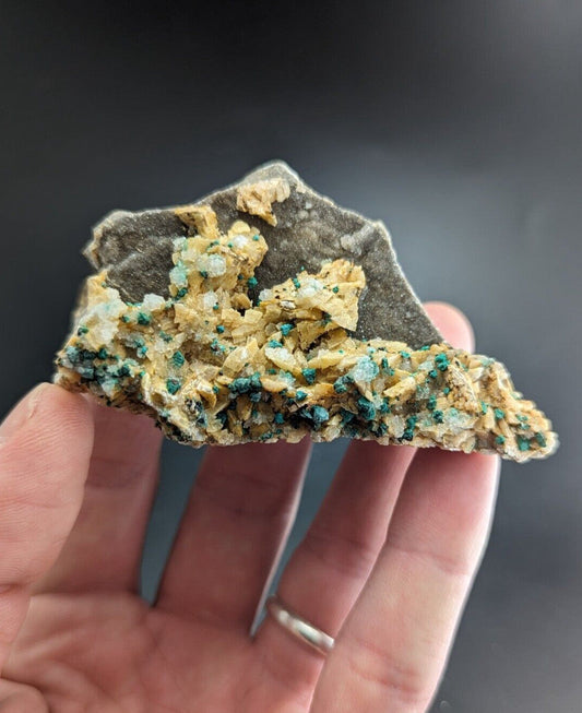 A hand holds a mineral specimen featuring vibrant green crystals growing from a rough, grayish matrix with golden-yellow crystalline formations.