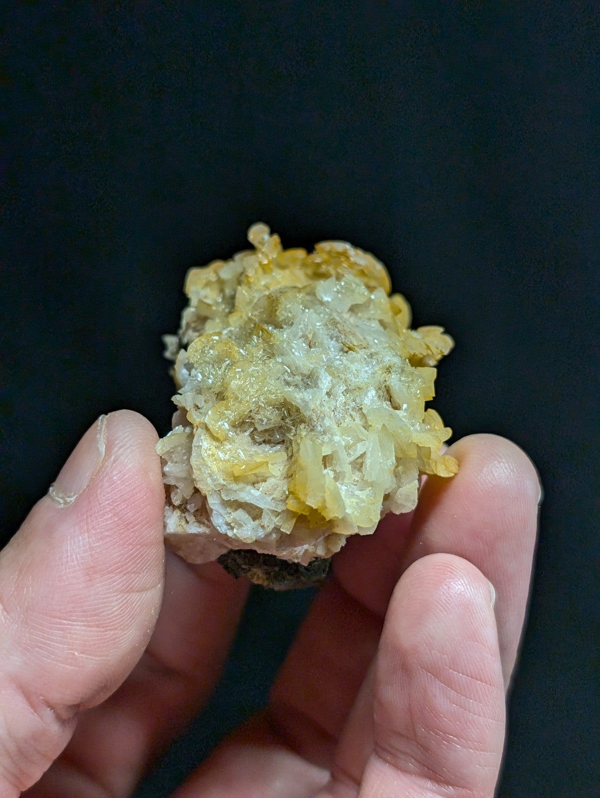 A hand holds a cluster of translucent yellow and white crystalline minerals against a dark background.
