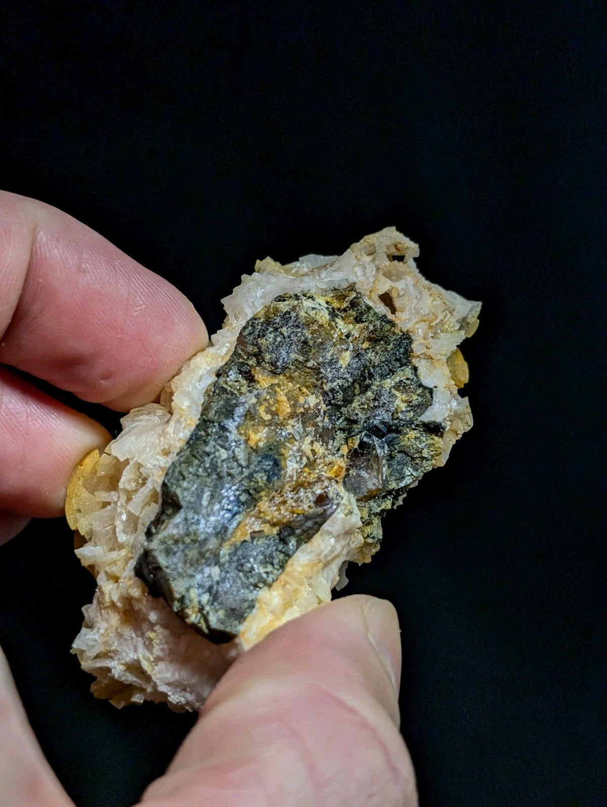 A hand holds a rough, irregularly shaped mineral specimen featuring dark green and yellowish hues embedded in translucent white crystalline matrix.
