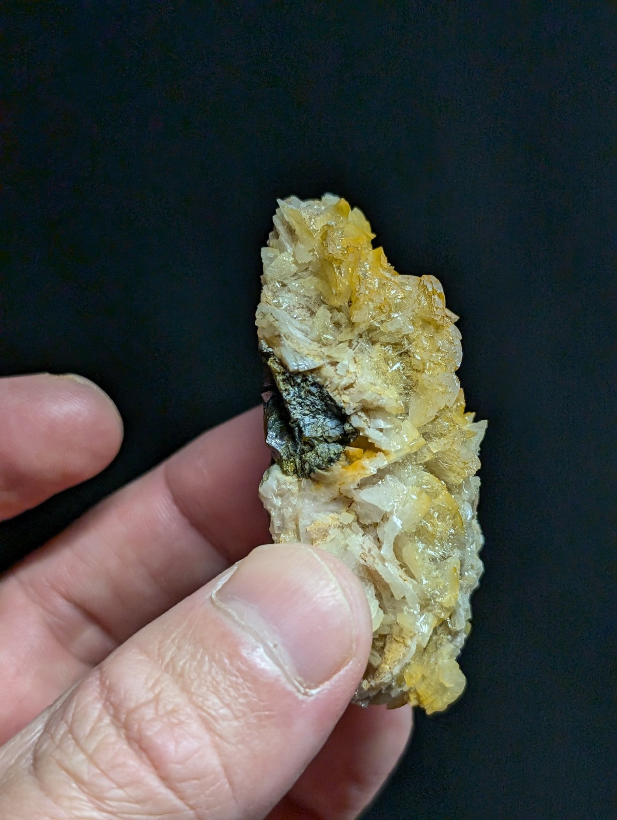 A hand holds a textured, irregularly shaped mineral specimen with patches of yellow and white crystalline formations.