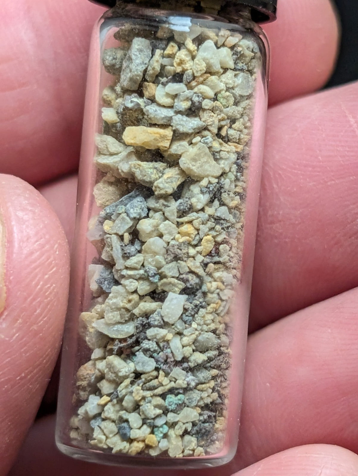 A clear glass vial filled with a mix of small, irregularly shaped rock fragments in shades of gray, beige, and yellow.