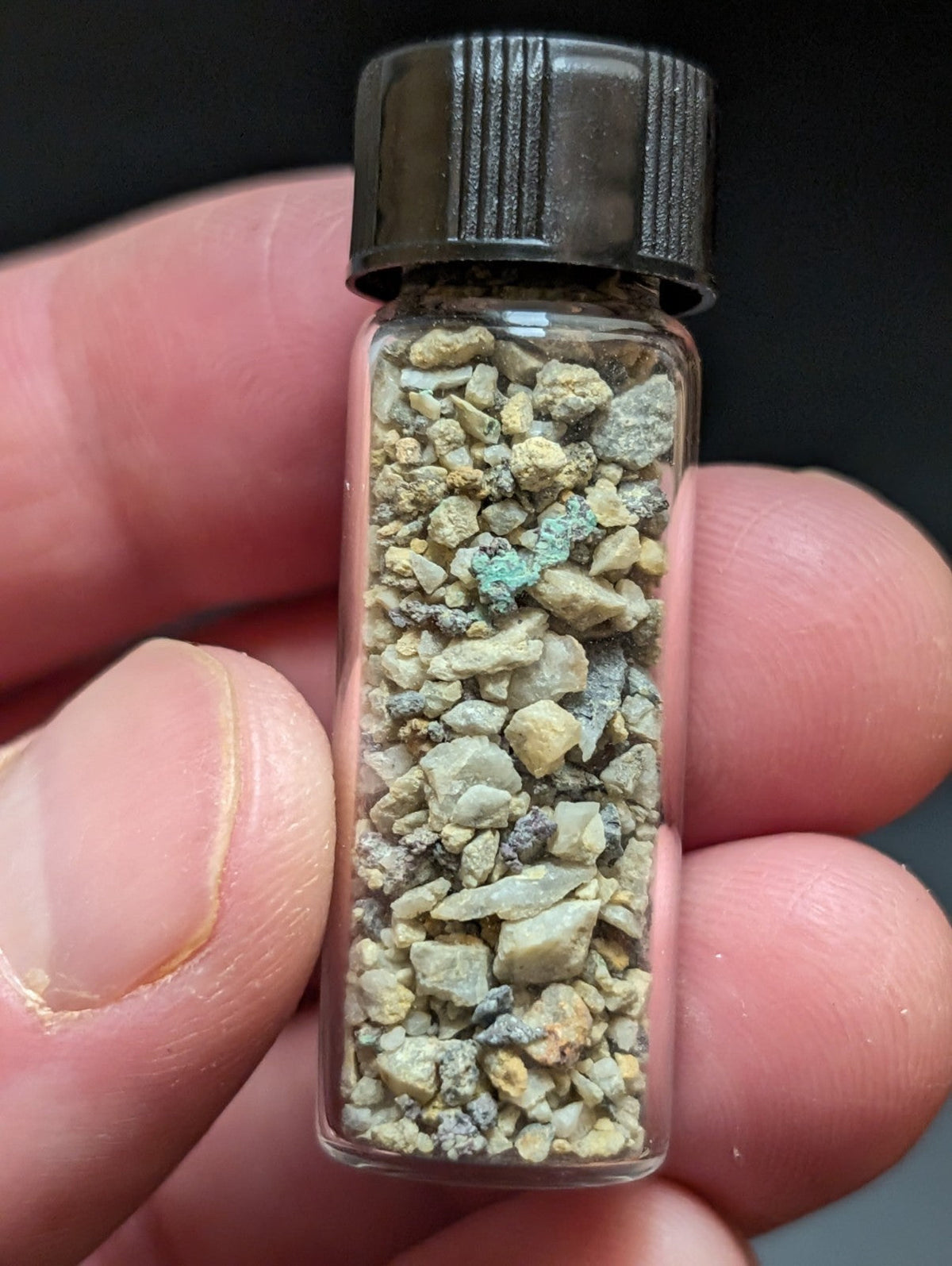 A clear glass vial with a black ribbed cap contains a mixture of small, irregularly shaped mineral fragments in shades of beige, gray, and green.
