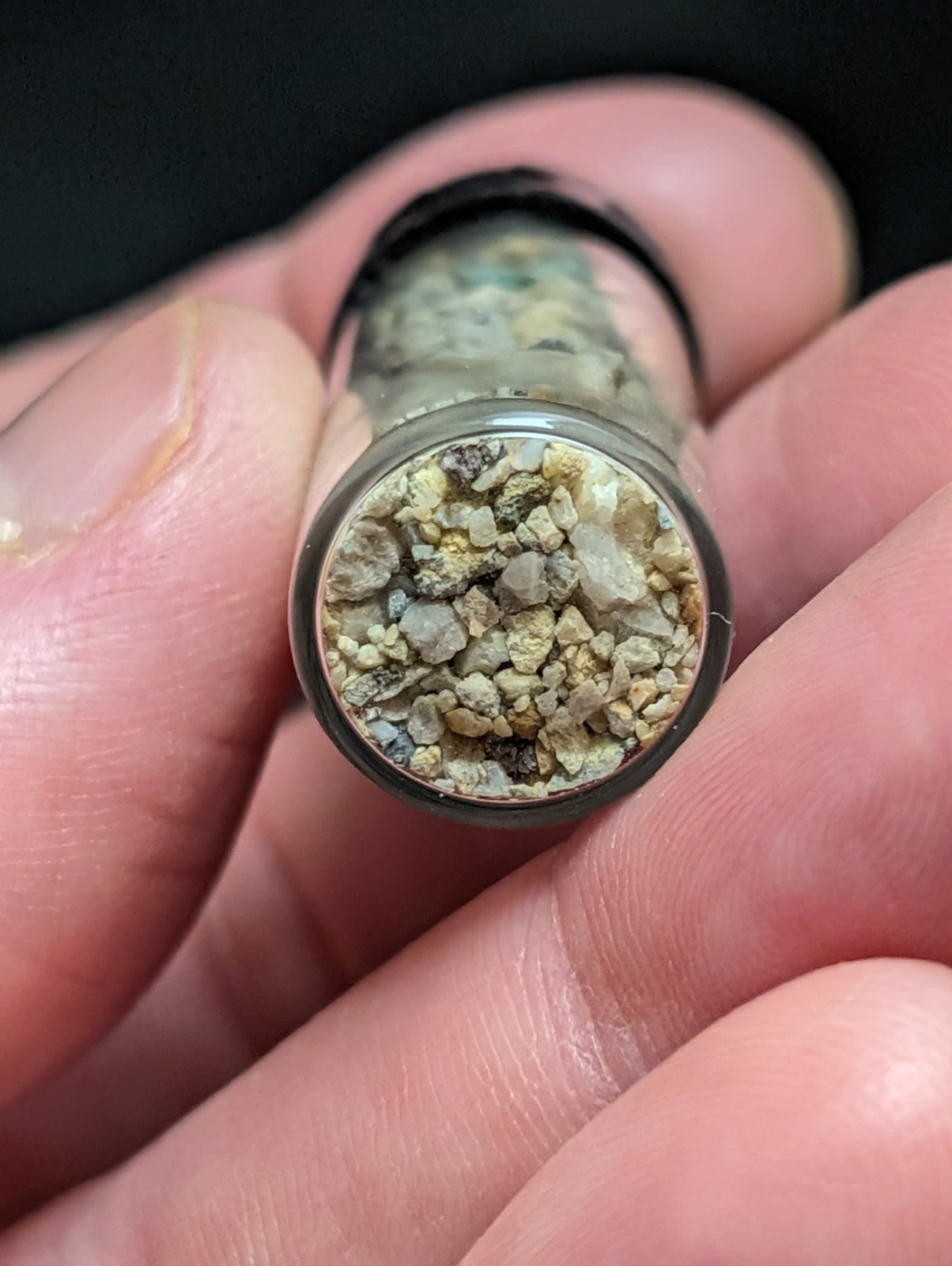 A clear glass vial filled with a mixture of small, irregularly shaped pebbles and mineral fragments in shades of beige, gray, and faint green.