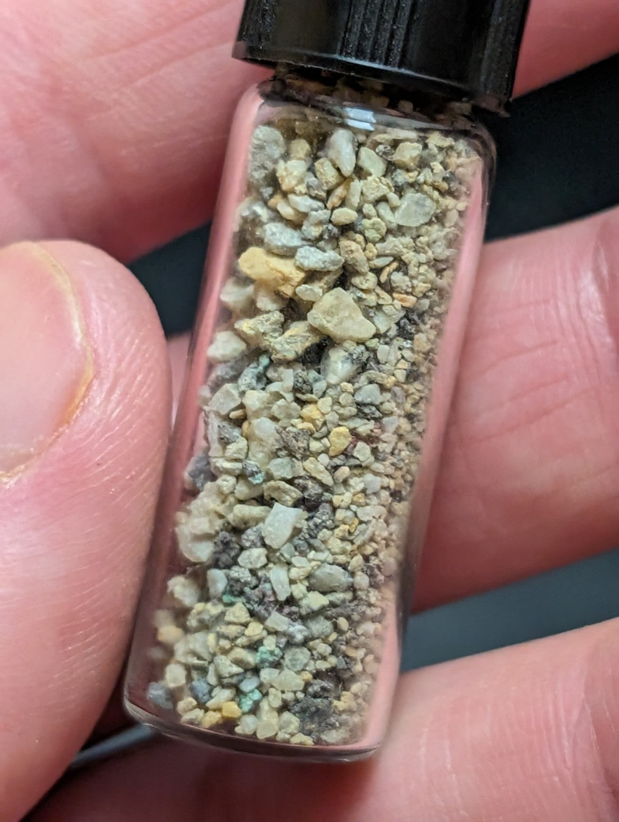A clear glass vial with a black ribbed cap contains a mixture of small, irregularly shaped fragments in shades of gray, green, and yellow.
