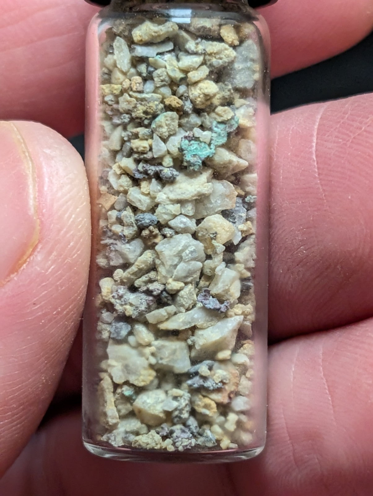 A clear glass vial filled with a mixture of small, irregularly shaped mineral fragments in shades of beige, gray, and patches of green.