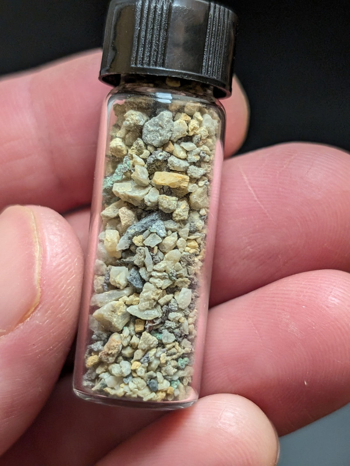 A clear glass vial with a black ribbed cap contains a mixture of small, irregularly shaped mineral fragments in shades of yellow, gray, and green.