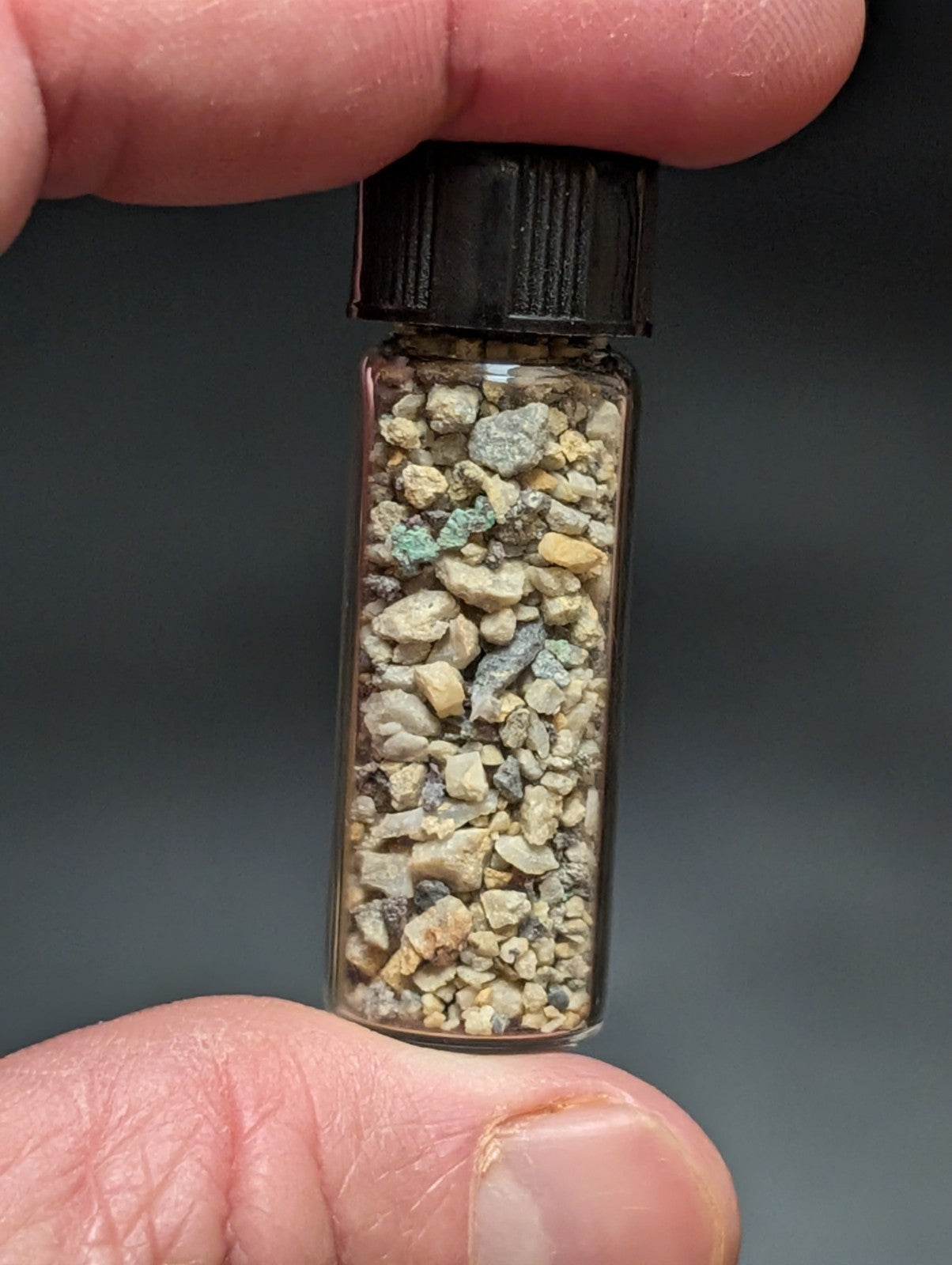 A clear glass vial with a black ribbed cap contains a mixture of small, irregularly shaped rock fragments in shades of beige, gray, and hints of green.