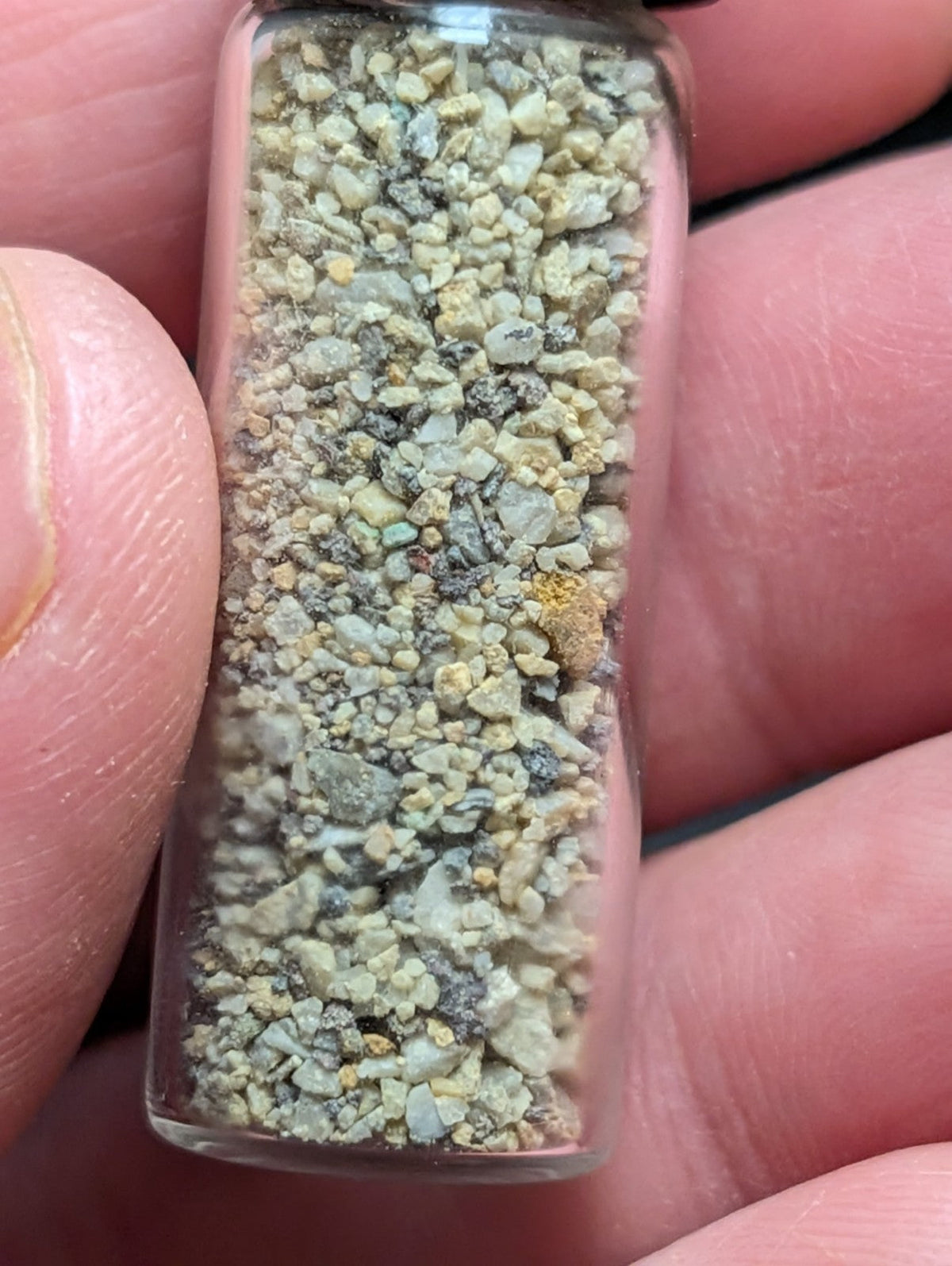A clear glass vial filled with a mixture of small, irregularly shaped gravel pieces in shades of gray, beige, and specks of yellow.