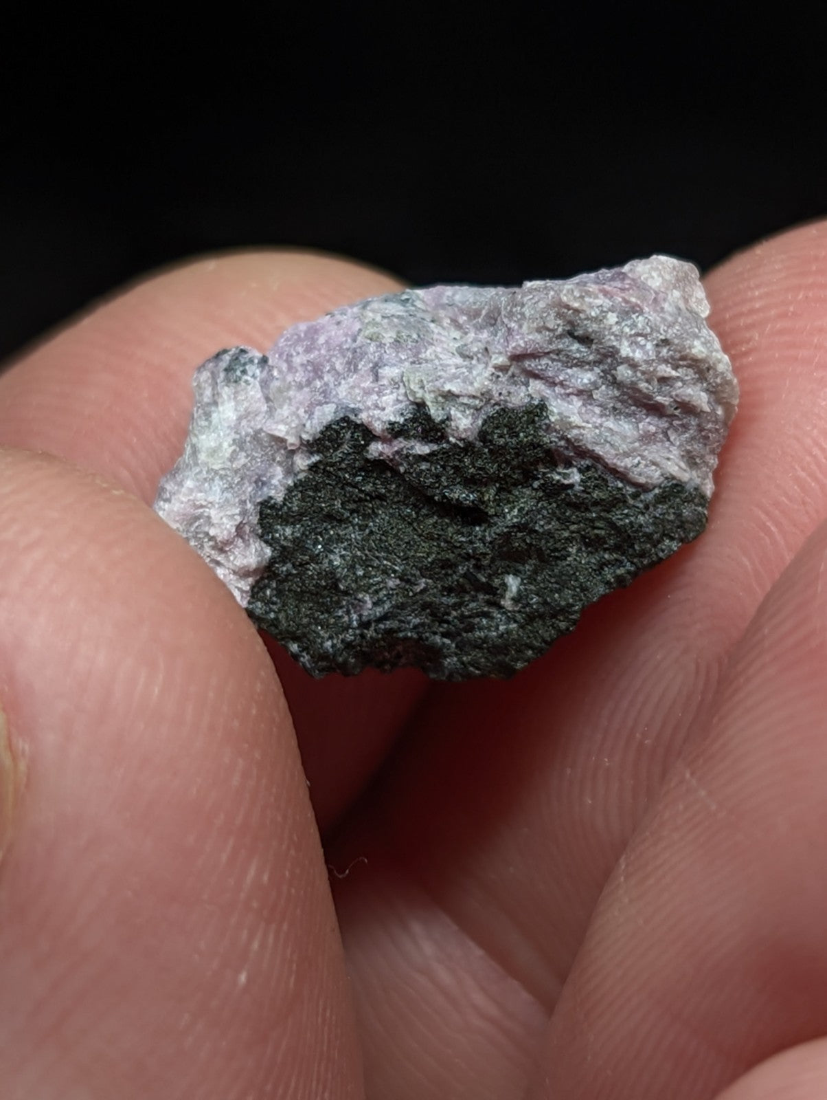 A rough, irregularly shaped mineral specimen with a mottled surface of grayish-purple and dark green hues.