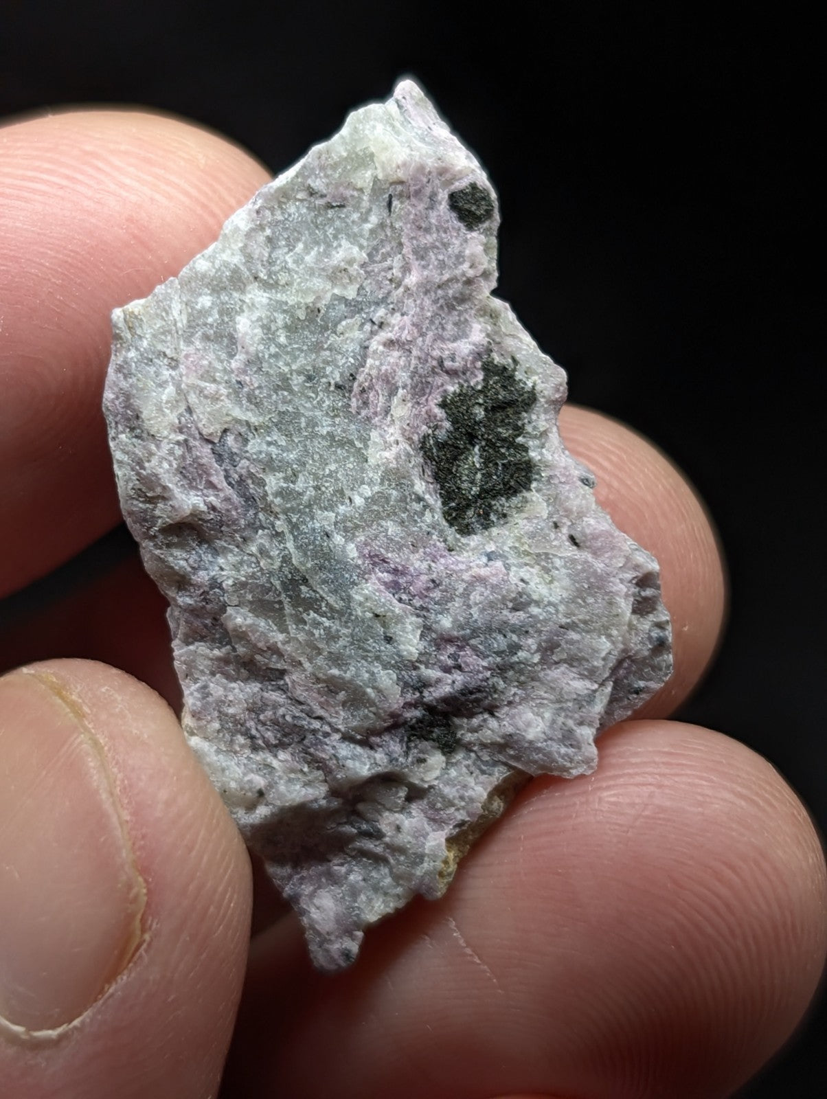 A rough, irregularly shaped mineral specimen with a mottled gray and purple surface featuring dark green inclusions.