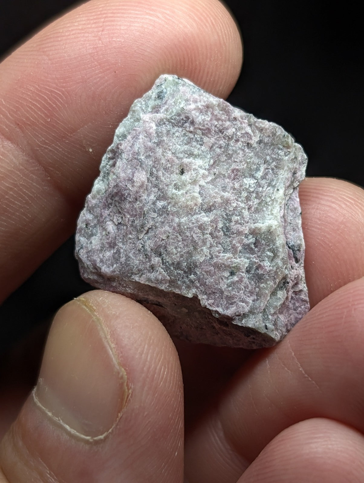 A rough, irregularly shaped stone with a mottled green and gray surface held gently between fingers.