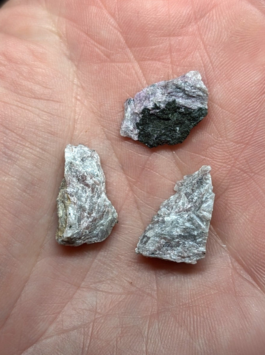 Three irregularly shaped, rough-textured mineral fragments with a mix of gray and dark green hues rest in an open palm.