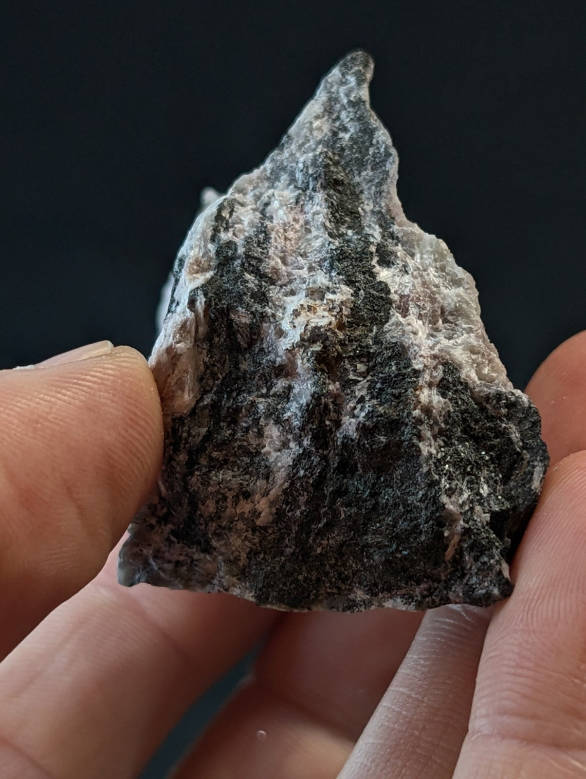 A rugged, irregularly shaped rock with a mottled surface of dark gray and speckles of pinkish-white mineral.