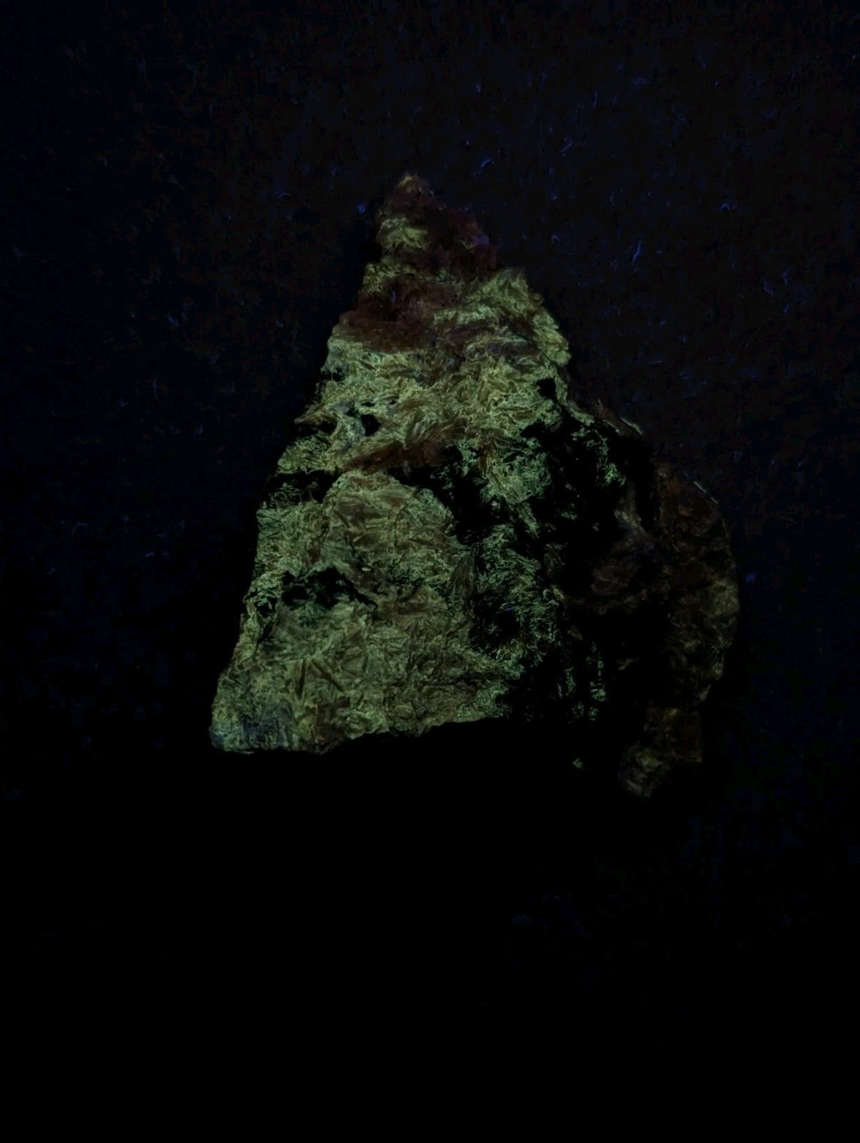 A rough, triangular-shaped mineral specimen with a textured surface displaying patches of greenish-yellow and dark brown hues.