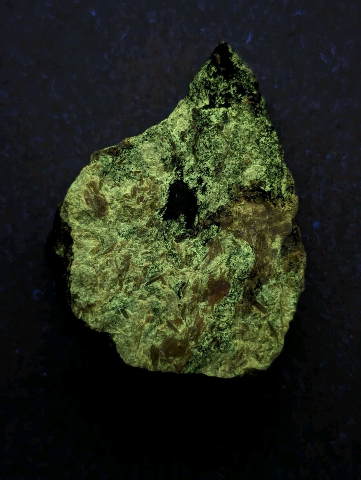 A rough, irregularly shaped green mineral specimen with a textured surface and patches of darker brownish hues.